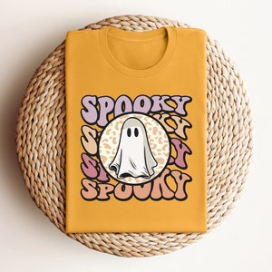 Puede incluir: Una camiseta amarilla mostaza con un fantasma blanco y el texto "Spooky" en letras rosas y moradas.