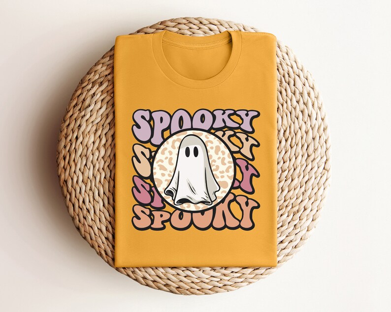 Puede incluir: Una camiseta amarilla mostaza con un fantasma blanco y el texto "Spooky" en letras rosas y moradas.