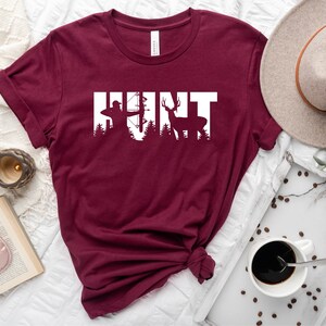 May include: Maroon t-shirt with the word "HUNT" in white text. The letters are formed by silhouettes of a hunter with a bow and arrow, a tree, and a deer.