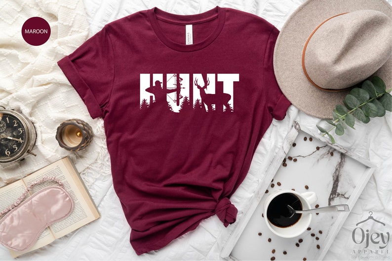May include: Maroon t-shirt with the word "HUNT" in white text. The letters are formed by silhouettes of a hunter with a bow and arrow, a tree, and a deer.