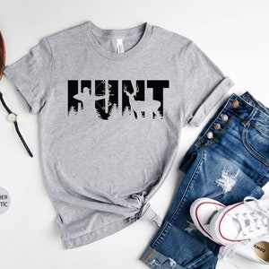 May include: A heather gray t-shirt with the word "HUNT" printed in black. The letters are formed by silhouettes of a hunter with a bow and arrow and a deer.