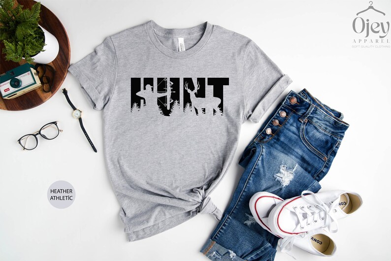 May include: A heather gray t-shirt with the word "HUNT" printed in black. The letters are formed by silhouettes of a hunter with a bow and arrow and a deer.