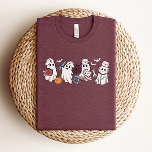 May include: A maroon t-shirt featuring a graphic of four ghosts dressed as nurses. The ghosts are holding medical supplies and a cauldron. The text "Boo Crew" is printed on the shirt.
