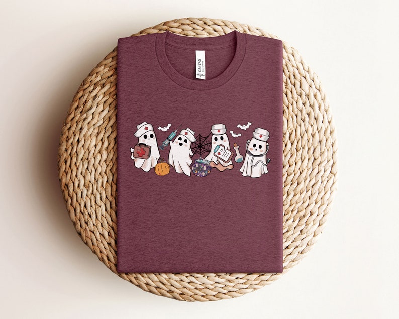 May include: A maroon t-shirt featuring a graphic of four ghosts dressed as nurses. The ghosts are holding medical supplies and a cauldron. The text "Boo Crew" is printed on the shirt.