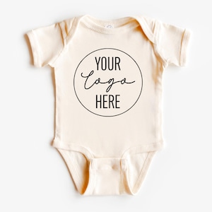 May include: A cream-colored baby onesie with a black circle and the text "YOUR LOGO HERE" in a handwritten font.
