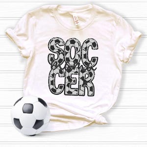 May include: White t-shirt with a black and white graphic that says "Soccer mama" with a soccer ball pattern.