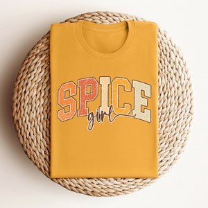 May include: A mustard yellow t-shirt with the text "SPICE girl" in a retro font with orange, red, and brown colors.