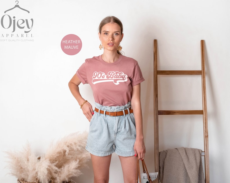 Op de afbeelding: Een heide-paarse t-shirt met een witte grafische afbeelding waarop "90's Baby" staat in een retro-lettertype. Het model draagt het shirt met een lichtblauwe denim short en een bruine riem.
