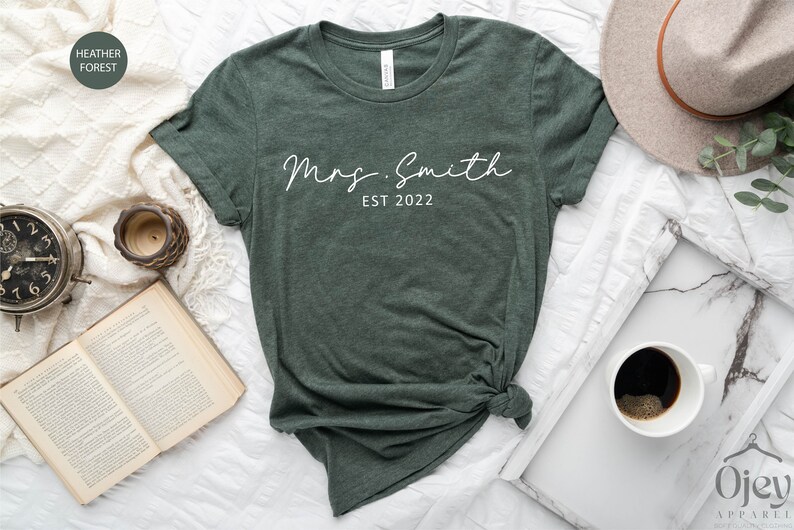 May include: A dark green t-shirt with the text "Mrs. Smith EST 2022" in white script font.