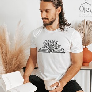 K&ouml;nnte beinhalten: Wei&szlig;es T-Shirt mit einer schwarzen Linienzeichnung eines offenen Buches, aus dem Blumen wachsen. Der Text "Ojay Apparel Soft Quality Clothing" befindet sich in der oberen rechten Ecke des Bildes.
