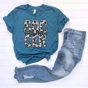 May include: Teal blue t-shirt with a white and black soccer ball design and the text "Soccer Mama" printed on the front. The shirt is paired with light blue jeans.