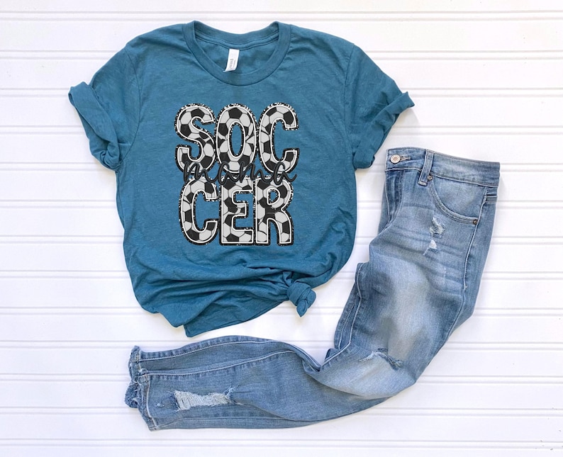 May include: Teal blue t-shirt with a white and black soccer ball design and the text "Soccer Mama" printed on the front. The shirt is paired with light blue jeans.