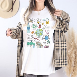 May include: White t-shirt with a colorful graphic design featuring illustrations of ways to help the planet. The graphic includes a bicycle, a water bottle, a recycling symbol, a faucet, a tree, a turtle, and the text "Save the planet, save a turtle, plant a tree, don't litter, save water, save life, recycle, reduce, reuse, turn off lights, skip a straw".