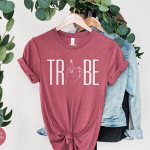 May include: A heather raspberry t-shirt with the word "TRIBE" printed in white. The "I" in "TRIBE" is replaced with a stylized image of a hand with a ring on the middle finger giving the middle finger.