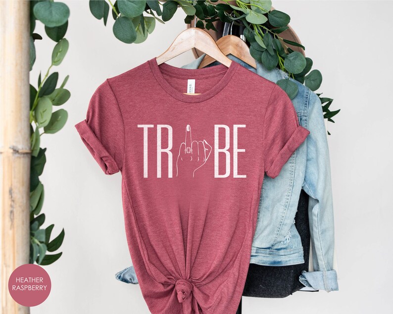 May include: A heather raspberry t-shirt with the word "TRIBE" printed in white. The "I" in "TRIBE" is replaced with a stylized image of a hand with a ring on the middle finger giving the middle finger.