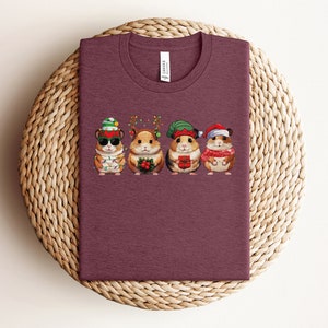 May include: A maroon t-shirt with a graphic of four hamsters wearing Christmas hats and holding gifts. The hamsters are wearing a Santa hat, an elf hat, a reindeer hat, and a Christmas tree hat.