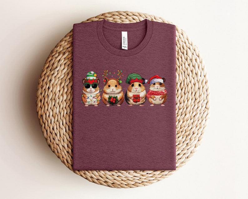 May include: A maroon t-shirt with a graphic of four hamsters wearing Christmas hats and holding gifts. The hamsters are wearing a Santa hat, an elf hat, a reindeer hat, and a Christmas tree hat.