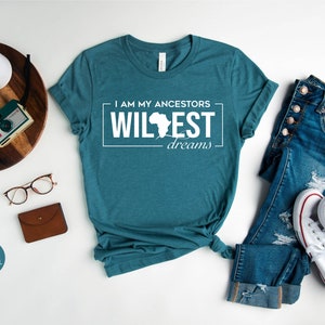 Peut inclure: Un t-shirt bleu sarcelle avec le texte "I am my ancestors' wildest dreams" et un contour du continent africain. Le t-shirt est pos&eacute; &agrave; plat sur une surface blanche avec un jean, une paire de baskets blanches, une montre, un appareil photo et une paire de lunettes.
