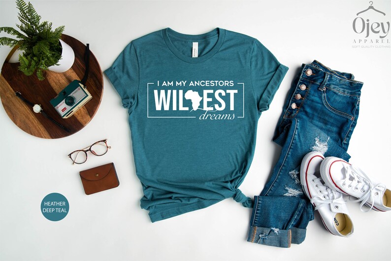 Peut inclure: Un t-shirt bleu sarcelle avec le texte "I am my ancestors' wildest dreams" et un contour du continent africain. Le t-shirt est pos&eacute; &agrave; plat sur une surface blanche avec un jean, une paire de baskets blanches, une montre, un appareil photo et une paire de lunettes.