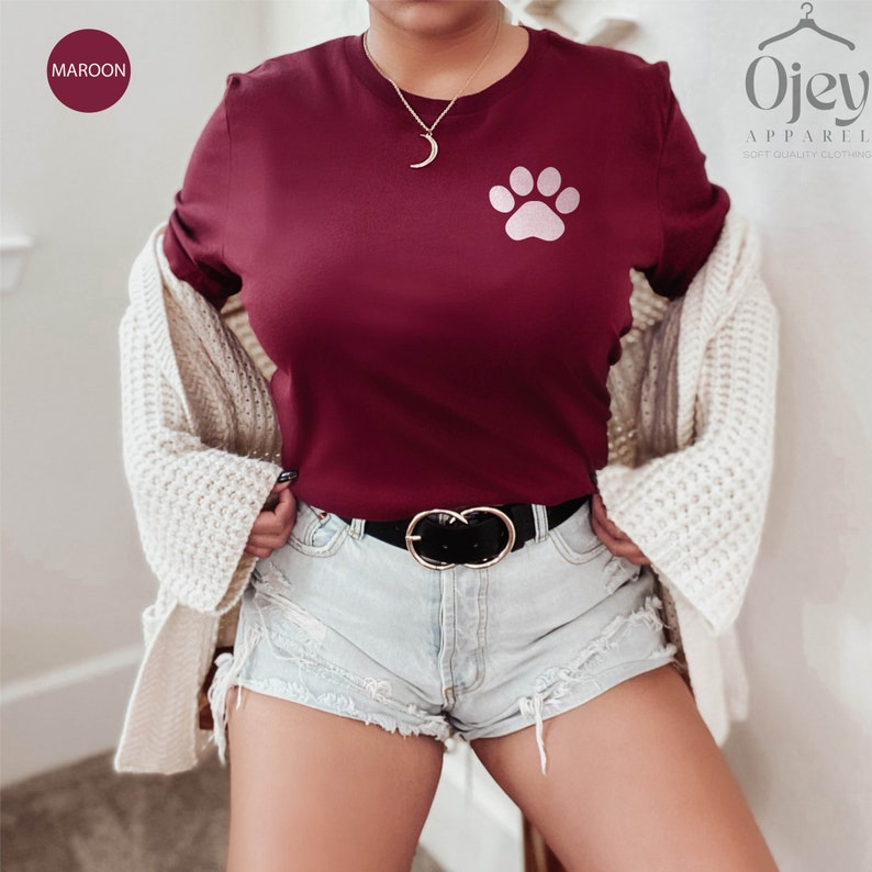 May include: A maroon t-shirt with a pink paw print graphic. The t-shirt is being worn by a person with a white cardigan and denim shorts.