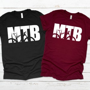 Puede incluir: Dos camisetas con la palabra "MTB" en letras blancas con una silueta de una bicicleta de monta&ntilde;a dentro de las letras. La camiseta de la izquierda es negra y la camiseta de la derecha es granate.