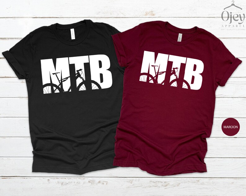 Puede incluir: Dos camisetas con la palabra "MTB" en letras blancas con una silueta de una bicicleta de monta&ntilde;a dentro de las letras. La camiseta de la izquierda es negra y la camiseta de la derecha es granate.