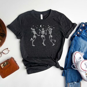 May include: Heather dark grey t-shirt featuring a graphic of three skeletons dancing under a crescent moon and stars. The t-shirt is displayed on a white background with a pair of jeans, a purse, and a pair of glasses.