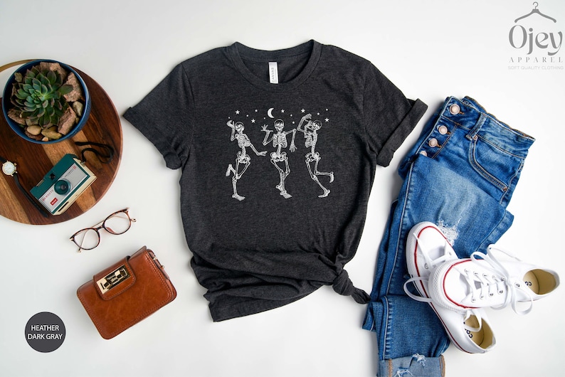 May include: Heather dark grey t-shirt featuring a graphic of three skeletons dancing under a crescent moon and stars. The t-shirt is displayed on a white background with a pair of jeans, a purse, and a pair of glasses.