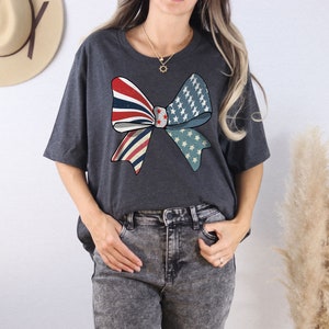 May include: A dark gray t-shirt with a large red, white, and blue bow design. The bow is made up of two overlapping bows, one with red and white stripes and the other with white stars on a blue background.
