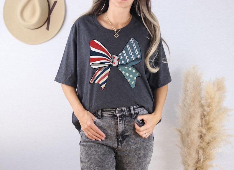 May include: A dark gray t-shirt with a large red, white, and blue bow design. The bow is made up of two overlapping bows, one with red and white stripes and the other with white stars on a blue background.