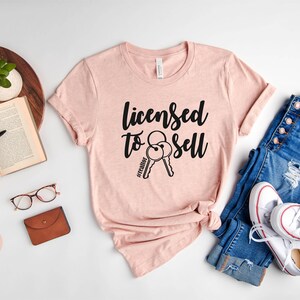 Pode incluir: Uma camiseta rosa com o texto "Licensed to Sell" e um gr&aacute;fico de duas chaves. A camiseta est&aacute; colocada sobre uma superf&iacute;cie branca com um par de cal&ccedil;as de ganga e t&eacute;nis brancos.