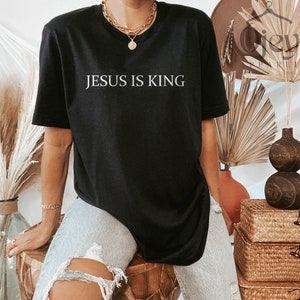 Pode incluir: Uma camiseta preta com o texto "JESUS IS KING" impresso em branco na frente. A camiseta est&aacute; sendo usada por uma pessoa com um colar de ouro.
