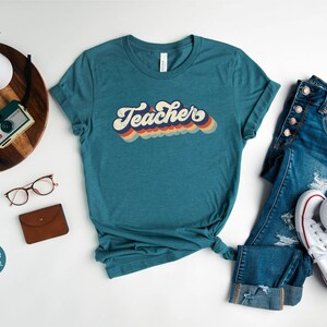 Puede incluir: Una camiseta azul turquesa con la palabra "Teacher" en una fuente retro arco&iacute;ris. La camiseta est&aacute; colocada sobre una superficie blanca con unas gafas, una cartera marr&oacute;n y una bandeja de madera con una c&aacute;mara y un reloj.
