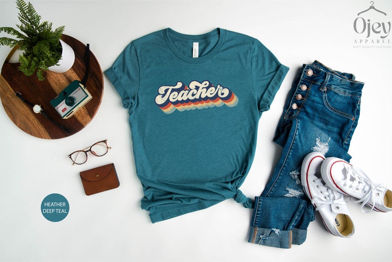 Puede incluir: Una camiseta azul turquesa con la palabra "Teacher" en una fuente retro arco&iacute;ris. La camiseta est&aacute; colocada sobre una superficie blanca con unas gafas, una cartera marr&oacute;n y una bandeja de madera con una c&aacute;mara y un reloj.