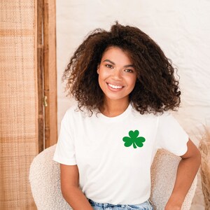 May include: A white t-shirt with a green shamrock design. The shirt is being worn by a person with curly hair. The person is sitting on a chair.