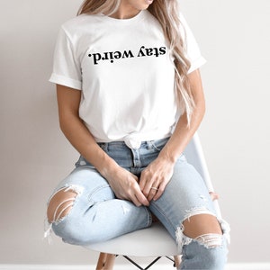 May include: A white t-shirt with the text "Stay Weird." printed in black. The shirt is being worn by a person sitting on a white chair. The person is wearing blue jeans with distressed details.