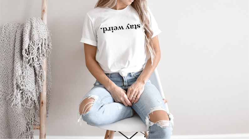 May include: A white t-shirt with the text "Stay Weird." printed in black. The shirt is being worn by a person sitting on a white chair. The person is wearing blue jeans with distressed details.