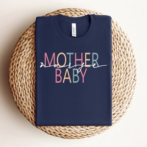 Puede incluir: Una camiseta azul marino con el texto "MOTHER of a BABY" en letras de colores. Las letras son de diferentes colores, incluyendo rosa, azul claro y amarillo.