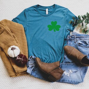 May include: A heather deep teal t-shirt with a green shamrock graphic. The shirt is displayed on a white surface with a brown sweater, blue jeans, and brown boots.