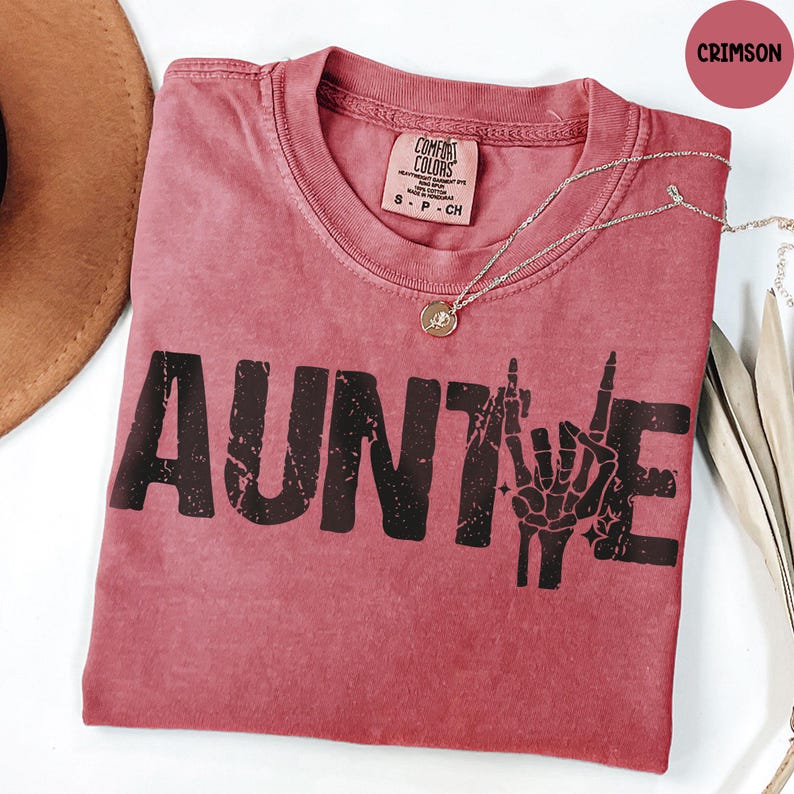 May include: Crimson-colored t-shirt with the word "AUNTIE" in distressed black lettering. The "E" is stylized with a skeleton hand gesture. A silver necklace with a pendant is draped across the shirt.