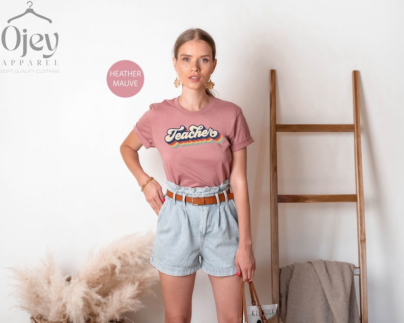 Puede incluir: Una camiseta de color malva jaspeado con el texto "Teacher's" en un dise&ntilde;o retro arco&iacute;ris.