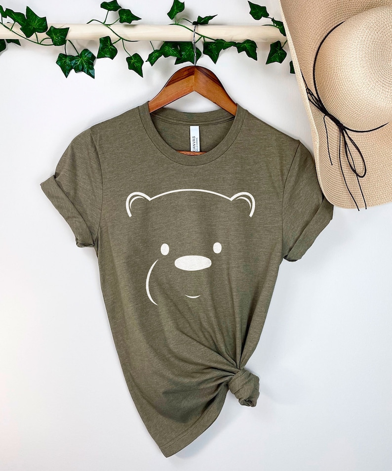 May include: A green t-shirt with a white bear face graphic. The shirt is knotted at the bottom and hanging on a wooden hanger.