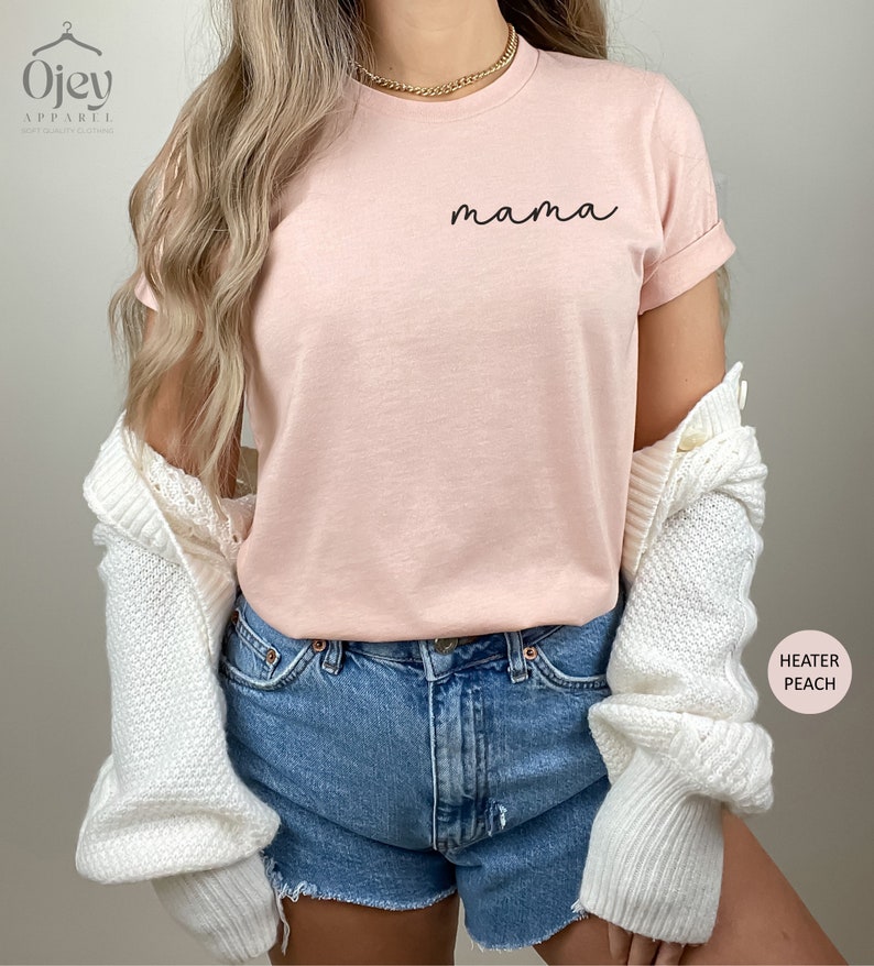 以下が含まれることがあります： ピンクのTシャツに、黒い筆記体で「mama」と書いてあります。Tシャツは、白いカーディガンと青いデニムのショートパンツを着用した人が着ています。