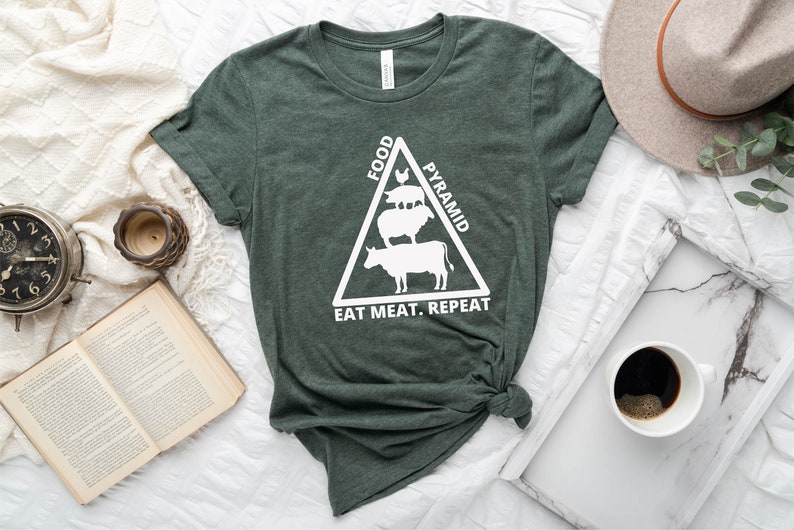May include: A green t-shirt with a white graphic of a food pyramid featuring a cow, sheep, pig, and chicken. The text "Food Pyramid" is written on the right side of the triangle and "Eat Meat. Repeat" is written below the triangle.