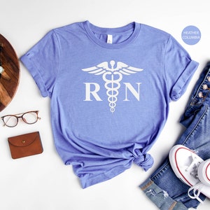 May include: A light blue t-shirt with a white medical caduceus symbol and the letters "RN" printed on the front. The shirt is folded and displayed on a white background.