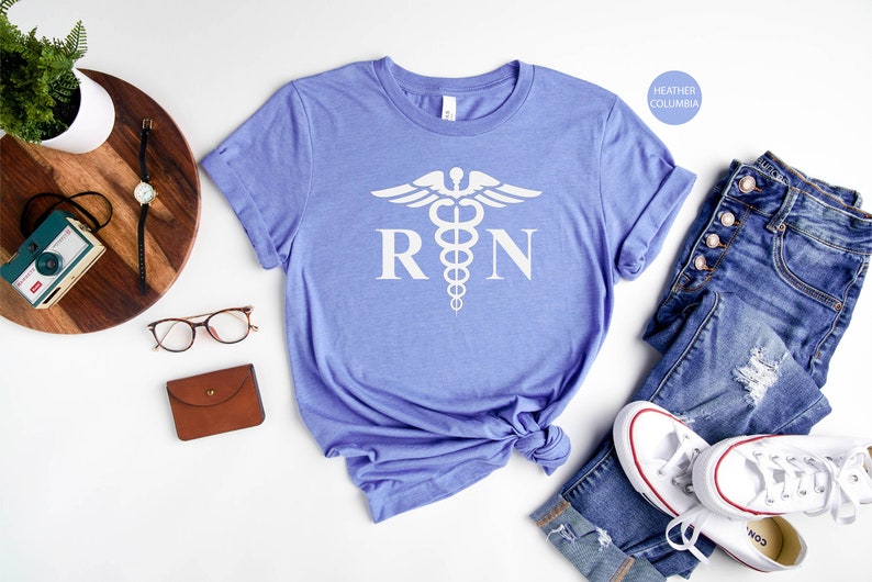 May include: A light blue t-shirt with a white medical caduceus symbol and the letters "RN" printed on the front. The shirt is folded and displayed on a white background.