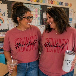 以下が含まれることがあります： お揃いの赤いTシャツを着た2人の女性。前面には「Redhawks Marshall」とプリントされています。一人はストロー付きの透明なプラスチックカップを持ち、もう一人は「teacher fuel」とプリントされた白いタンブラーを持っています。