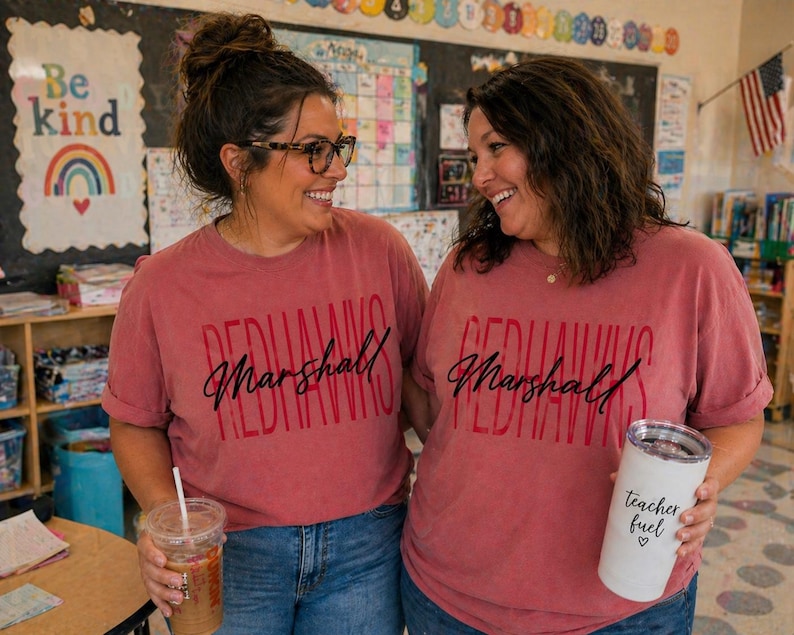 以下が含まれることがあります： お揃いの赤いTシャツを着た2人の女性。前面には「Redhawks Marshall」とプリントされています。一人はストロー付きの透明なプラスチックカップを持ち、もう一人は「teacher fuel」とプリントされた白いタンブラーを持っています。