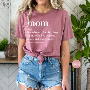 Może przedstawiać: R&oacute;żowy T-shirt z napisem "mom" zdefiniowanym jako "Osoba, kt&oacute;ra poświęca sw&oacute;j sen, zdrowie psychiczne, życie towarzyskie, pieniądze i kt&oacute;ra sika sama (patrz także: 'superwoman')".