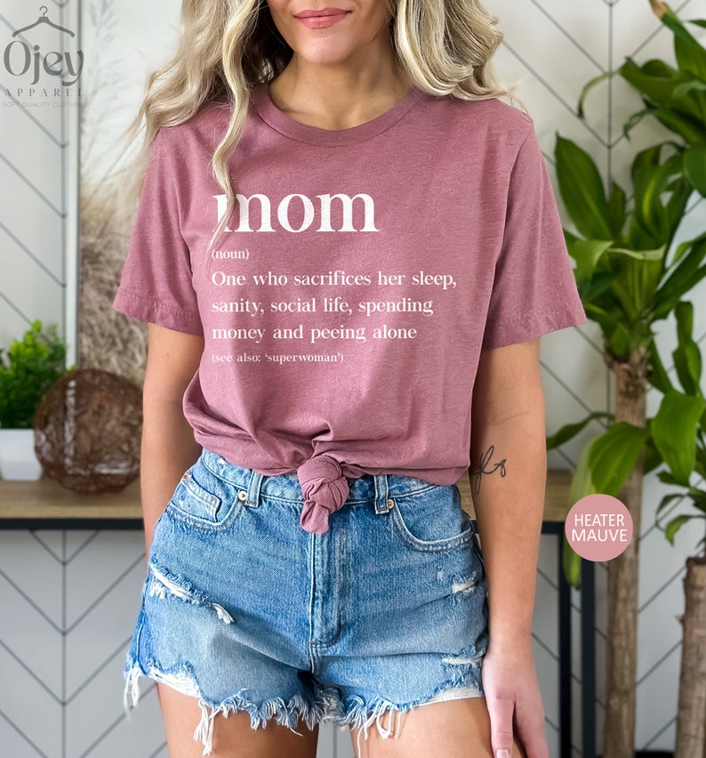 Może przedstawiać: R&oacute;żowy T-shirt z napisem "mom" zdefiniowanym jako "Osoba, kt&oacute;ra poświęca sw&oacute;j sen, zdrowie psychiczne, życie towarzyskie, pieniądze i kt&oacute;ra sika sama (patrz także: 'superwoman')".
